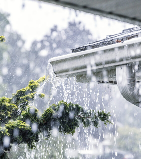 Drenching downpour rain storm water is overflowing off the tile shingle roof - streaming, rushing and splashing out over the overhanging eaves trough aluminum roof gutter system on a suburban residential colonial style house near Rochester, New York State, USA during a torrential mid-summer July downpour.